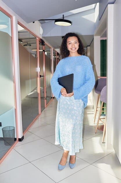 A woman is confidently standing in a modern office space, wearing a business casual outfit consisting of black trousers, a white button-down shirt, a black blazer, and stylish black loafers. She is holding a tablet and smiling slightly.