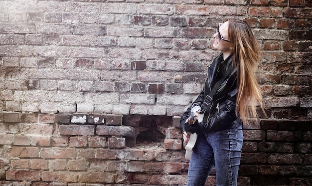 A woman is wearing a white t-shirt and blue jeans and is using a black leather jacket. This gives her look a vibe of a bad girl. The background is a brick wall filled with grafitti.