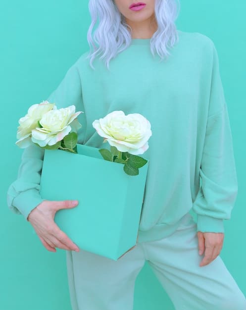 A woman wearing a head-to-toe teal outfit. She is wearing a teal sweater, teal trousers, and teal sneakers. She is holding a white clutch. The background is a modern, minimalist apartment.