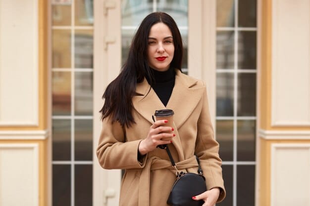 A woman wearing a beige trench coat, a white t-shirt, and black jeans. She is carrying a bright yellow tote bag. The background is a coffee shop.