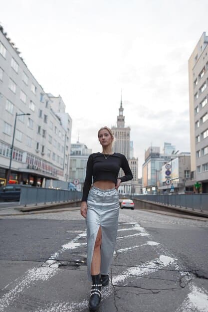 A woman walking down a city street wearing black leggings, a fitted black long-sleeve top, a stylish bomber jacket, and white sneakers. She is carrying a sleek black backpack. The background is a blurred cityscape with modern buildings.