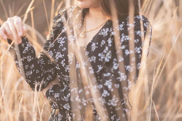 A close-up of a woman wearing a maxi dress with a delicate floral print, accessorized with a thin gold belt and layered necklaces. The background is a blurred outdoor setting.