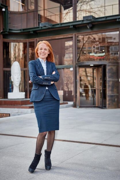 A woman wearing a belted blazer dress in a classic navy color, accessorized with a wide leather belt, pointed-toe heels, and a structured handbag, posing confidently in a professional setting.