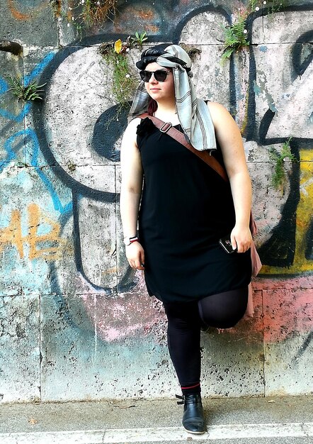 A woman standing in front of a colorful mural, wearing a fitted black athleisure dress with white sneakers and a denim jacket tied around her waist. She is wearing sunglasses and a baseball cap.