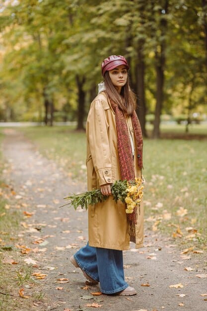 A woman wearing an autumn outfit consisting of an oversized sweater, corduroy pants, ankle boots, and a colorful scarf. She is walking down a street lined with trees in fall foliage.