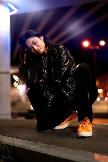 A woman posing in a dimly lit urban setting, wearing a delicate white lace top paired with a black leather jacket, skinny black jeans, and ankle boots.